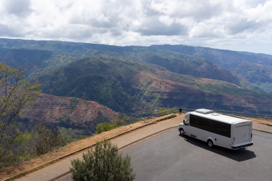 A tour coach parked at a Waimea Canyon lookout on Kauai with rust-red canyon walls behind
