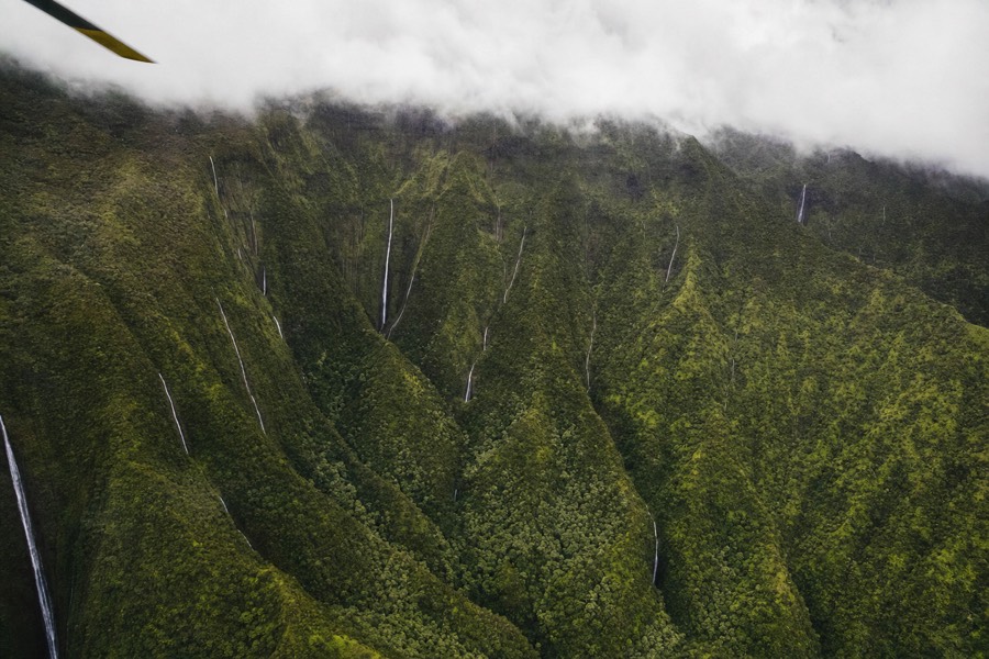 Helicopter in flight over green roadless valleys in Kauai's interior with waterfalls visible on the cliff walls
