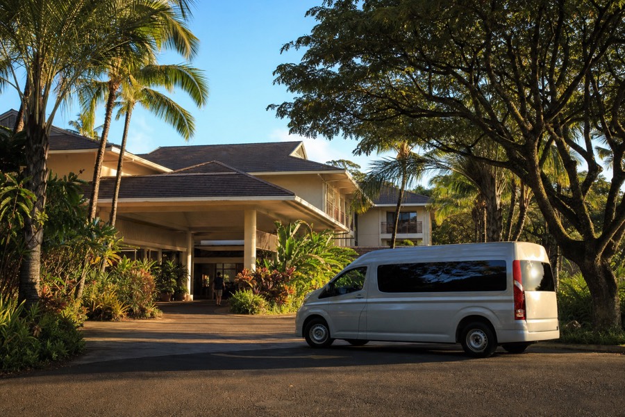 Small tour shuttle van parked outside a Kauai hotel entrance in early morning light, ready for hotel pickup