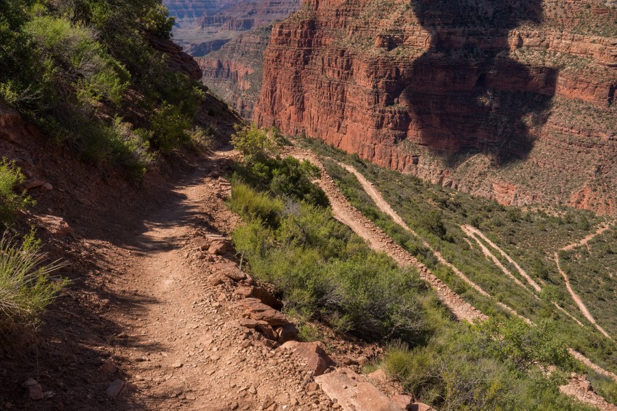 Trail descending into Waimea Canyon through red dirt and green vegetation, no hikers visible